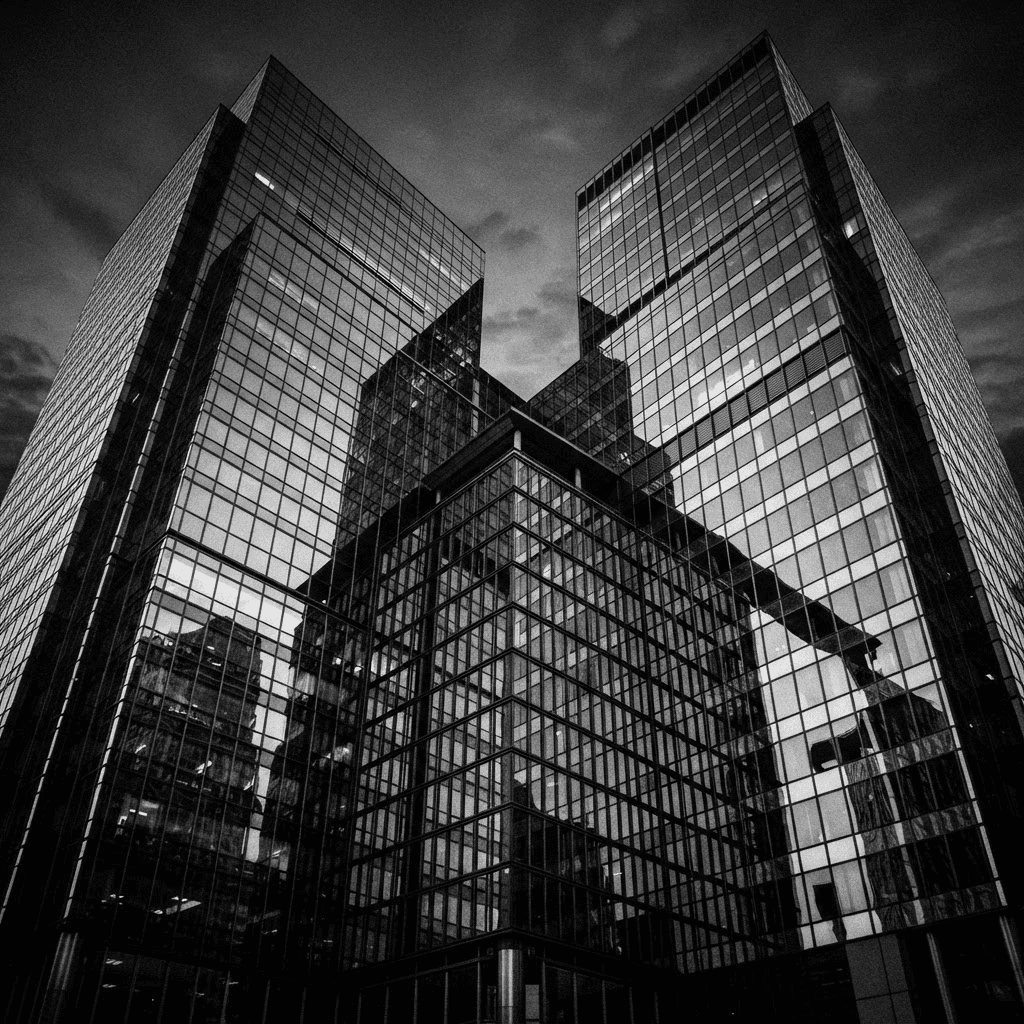 High-contrast black and white photograph of two overlapping glass office towers reflecting each other at dusk, suggesting a corporate merger